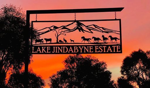 Silhouette of a metal sign reading "Lake Jindabyne Estate" against a vivid orange and pink sunset. The sign features cutouts of horses, cattle, and mountains, with tree silhouettes in the background