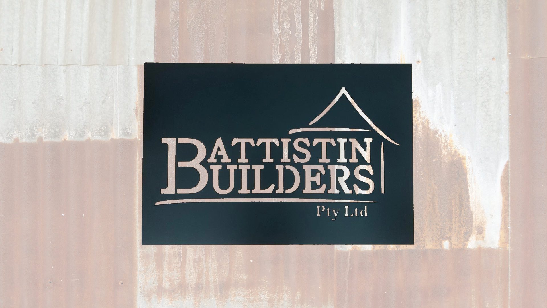 A sign mounted on a corrugated metal wall reads "Battistin Builders Pty Ltd" in bold white text. The word "Builders" features a roofline graphic extending from its top right corner, suggesting a house structure. The overall design is simple and professional.