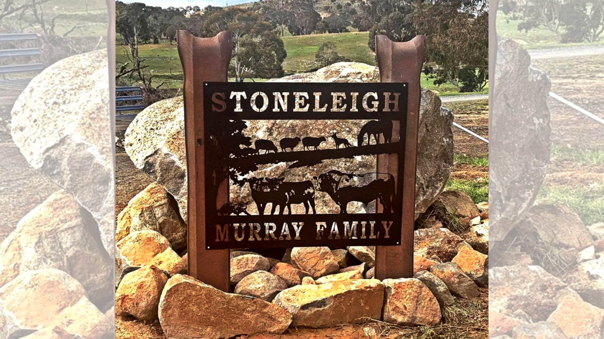 A metal sign reading "Stoneleigh Murray Family" is mounted on large rocks. The sign features cut-out silhouettes of livestock, including cows and horses. It is set against a rural landscape with trees and a grassy field in the background.