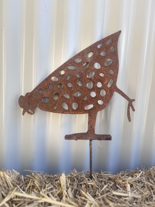 Rustic metal silhouette of a chicken with cut-out patterns, standing on straw against a corrugated background.