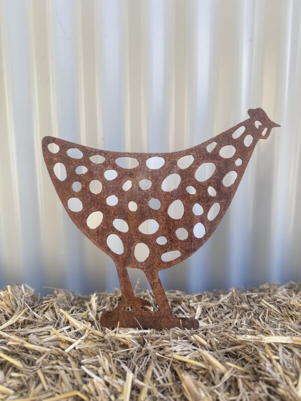 Rusted metal chicken silhouette with cut-out circles standing on straw against a corrugated wall.