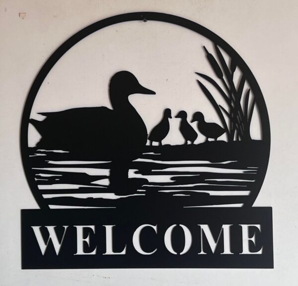 Black silhouette welcome sign featuring a duck in water with ducklings and reeds.