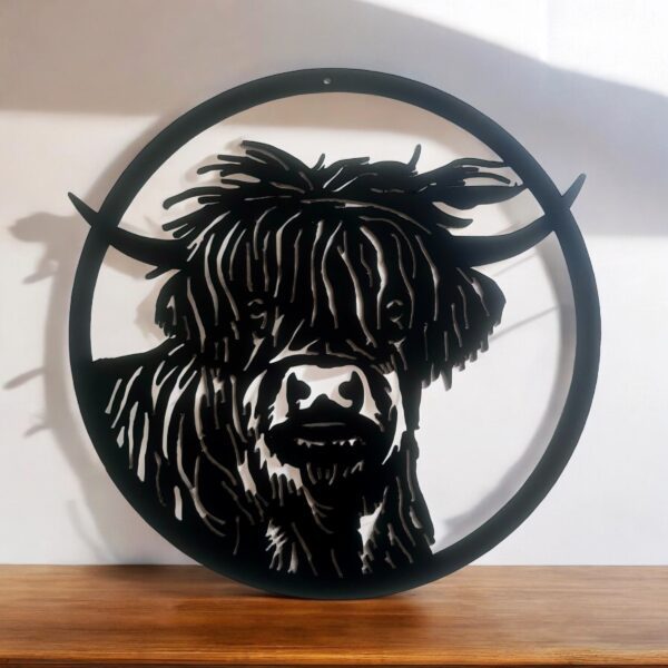 A circular metal wall art of a Highland cow on a wooden surface with shadows cast on a wall.