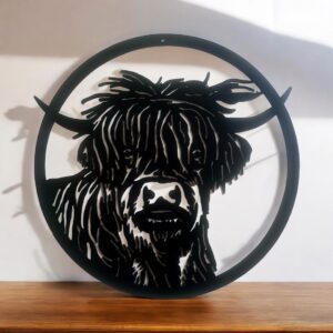 A circular metal wall art of a Highland cow on a wooden surface with shadows cast on a wall.