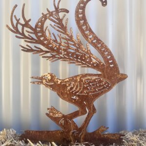 A rusted metal cutout of a peacock, with intricate details, standing on straw against a corrugated background.