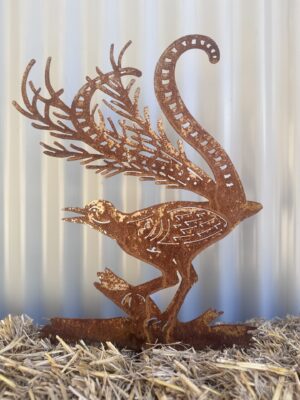 A rusted metal cutout of a peacock, with intricate details, standing on straw against a corrugated background.