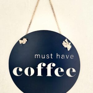 A round, navy blue sign with "must have coffee" in white letters, hung by a twine rope.