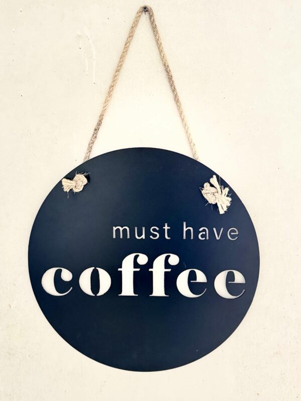 A round, navy blue sign with "must have coffee" in white letters, hung by a twine rope.