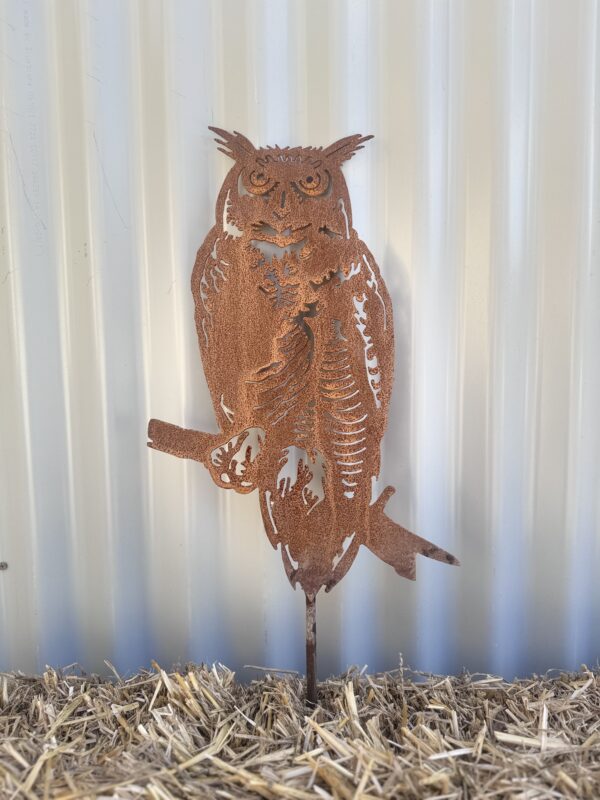 Rustic metal owl silhouette against a corrugated wall, mounted on straw-covered ground.