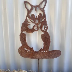 Rusty metal cutout of a fox licking its paw, mounted on a straw surface against a corrugated wall.