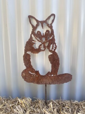 Rusty metal cutout of a fox licking its paw, mounted on a straw surface against a corrugated wall.