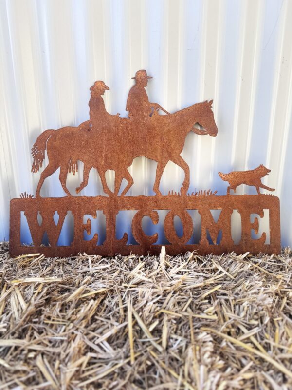 Rustic metal "WELCOME" sign with horse, rider, and dog silhouettes on straw, against a corrugated wall.