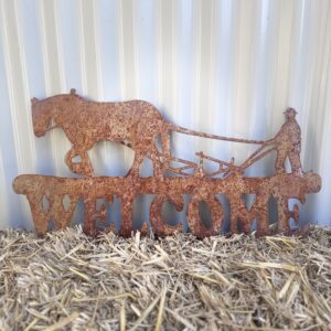 Rustic metal "WELCOME" sign with cut-out horses and a cowboy on straw against a corrugated wall.