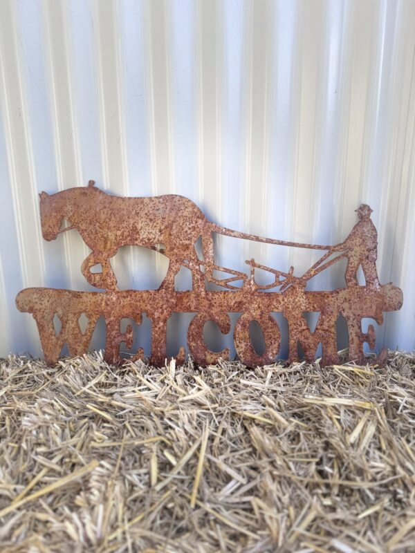 Rustic metal "WELCOME" sign with cut-out horses and a cowboy on straw against a corrugated wall.