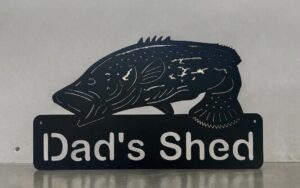 Metal sign with silhouette of a fish and the words "Dad's Shed" against a gray background.