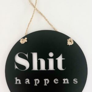 A circular black sign with white text "Shit happens" and a twine for hanging.