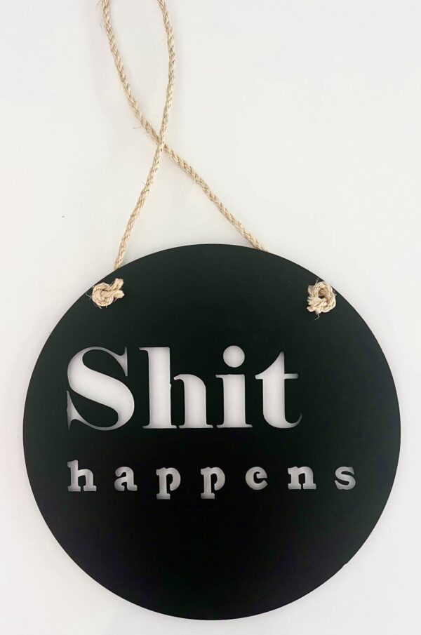 A circular black sign with white text "Shit happens" and a twine for hanging.