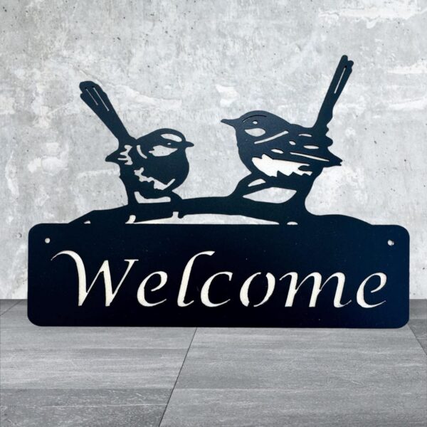 Metal "Welcome" sign with two birds perched, standing against a concrete wall.